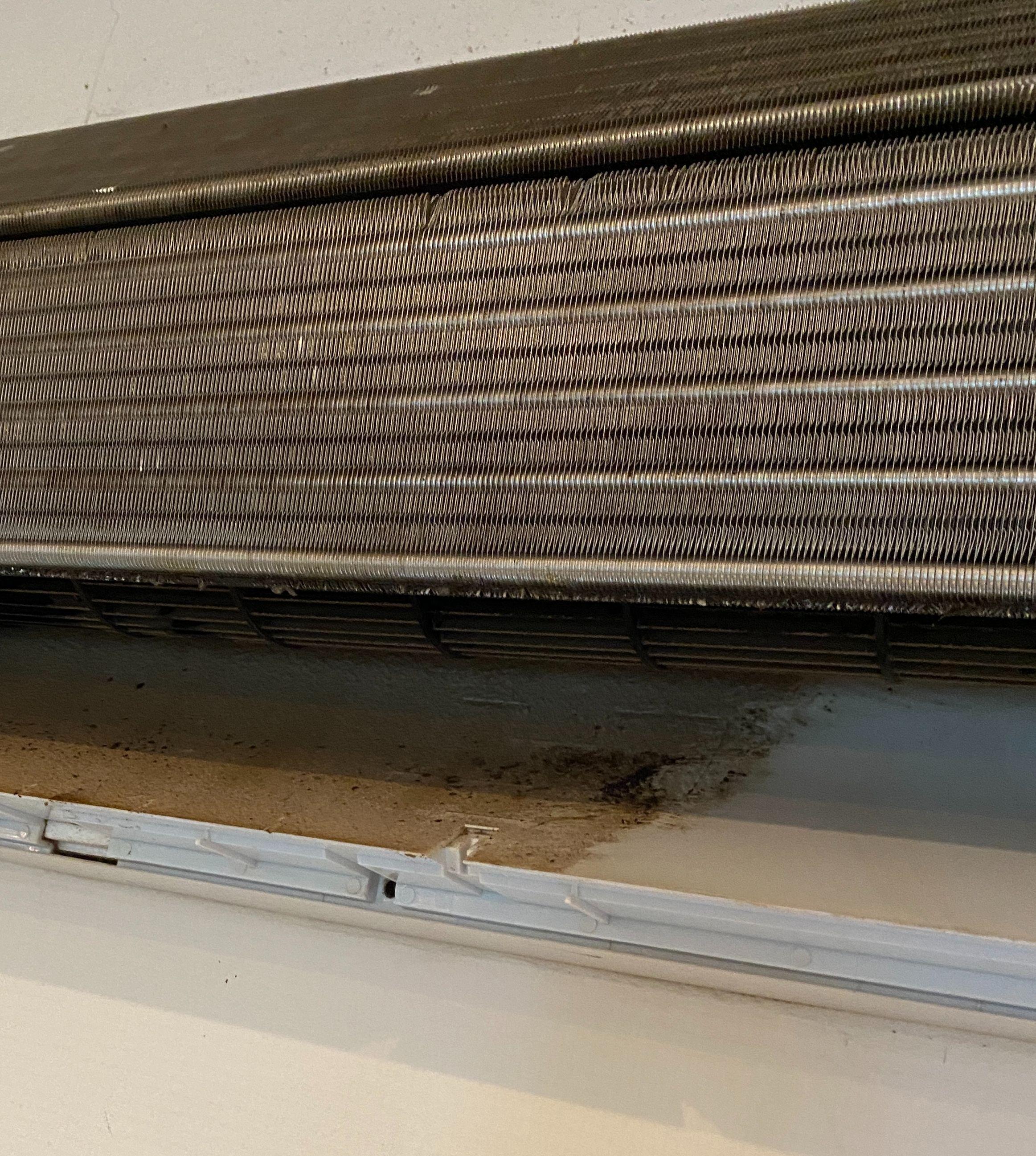Billede af en metalvarmeveksler med snavs og skimmel på væggen under den.