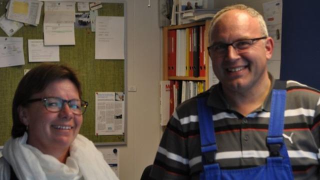 To personer smiler i et kontor, iført briller og arbejdstøj, omgivet af papirer og bøger.