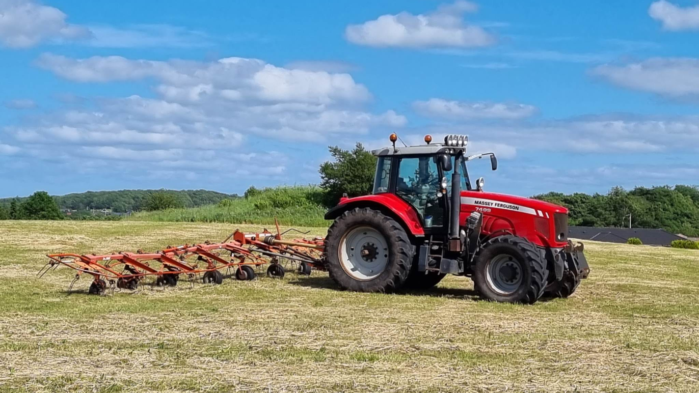 Rød traktor pløjer en mark under en blå himmel med skyer i baggrunden.