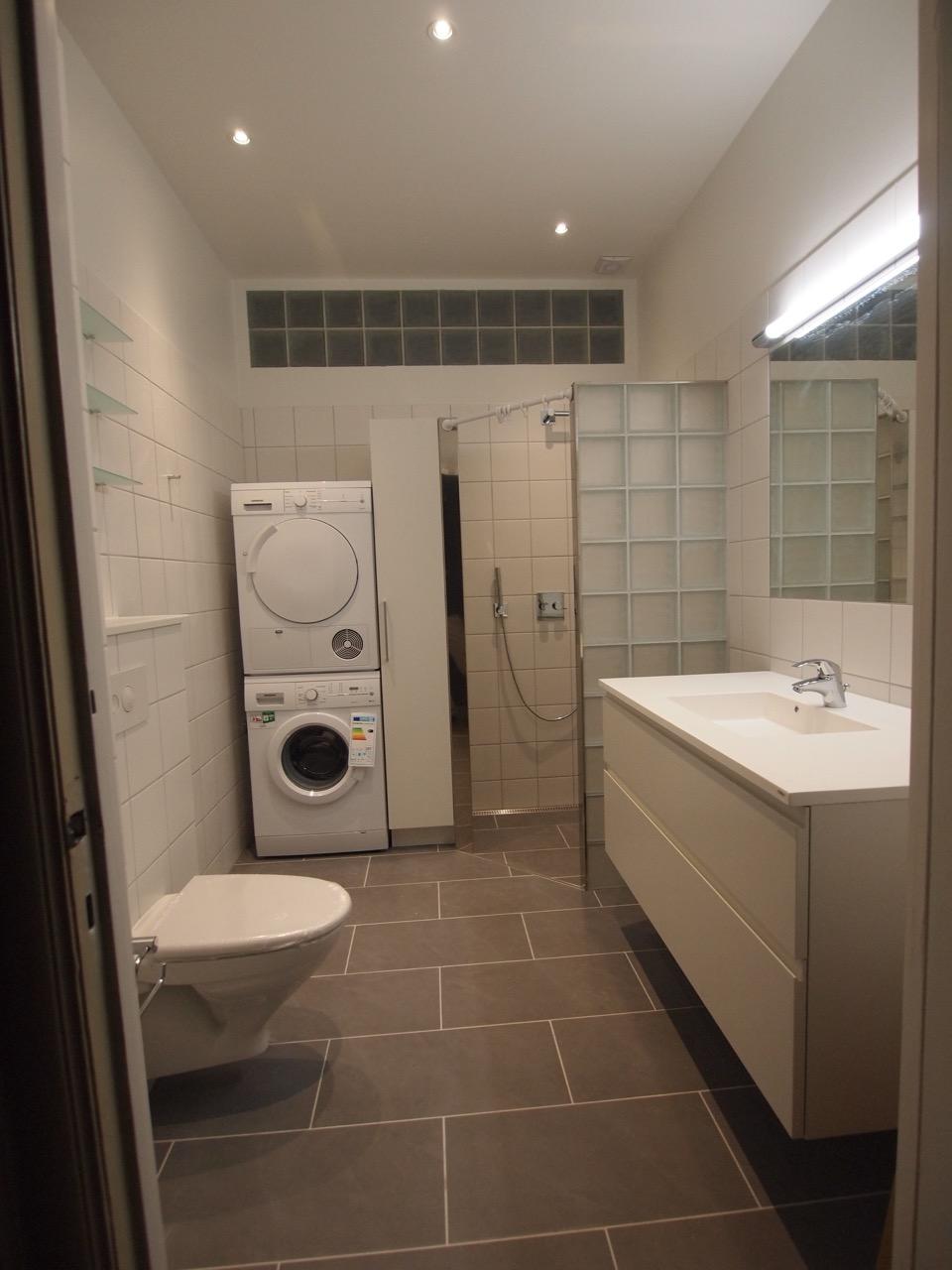 Moderne badeværelse med vaskemaskine, bruseniche, toilet og hvidt skab med håndvask.