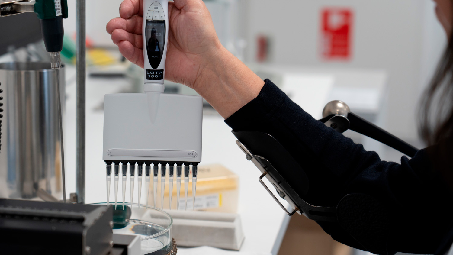En person bruger en pipette til at tage prøver i et laboratorium med forskellige laboratorieudstyr i baggrunden.