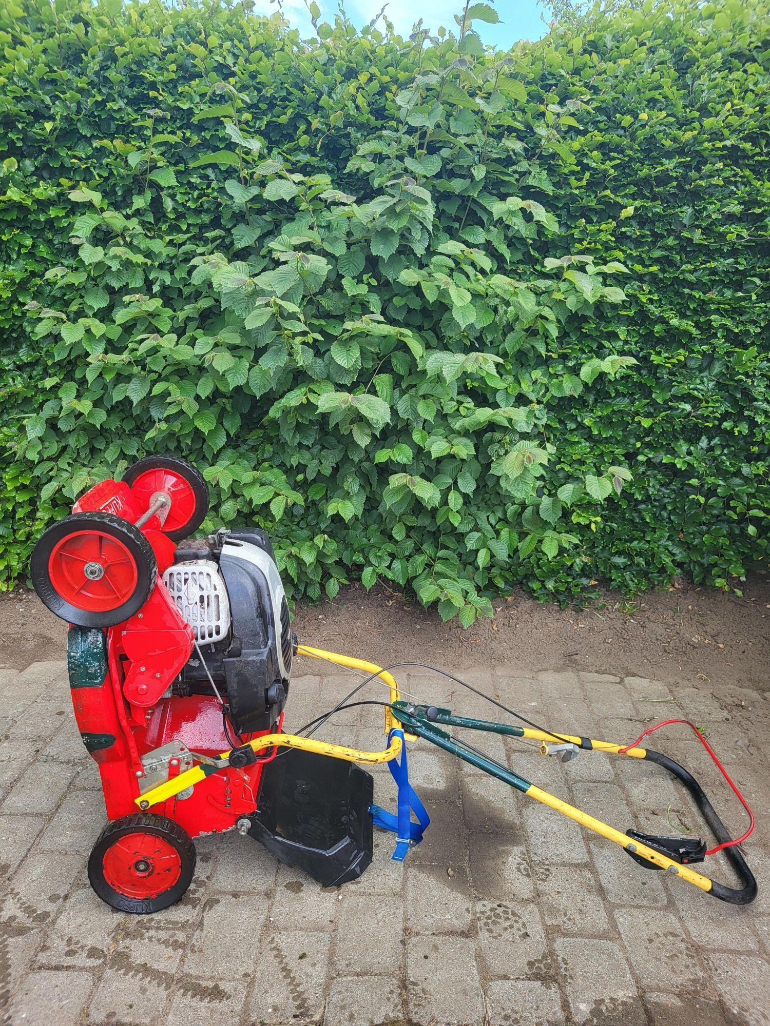 Rød plæneklipper med motor, der står på siden på en grusbelagt overflade ved en grøn hæk.