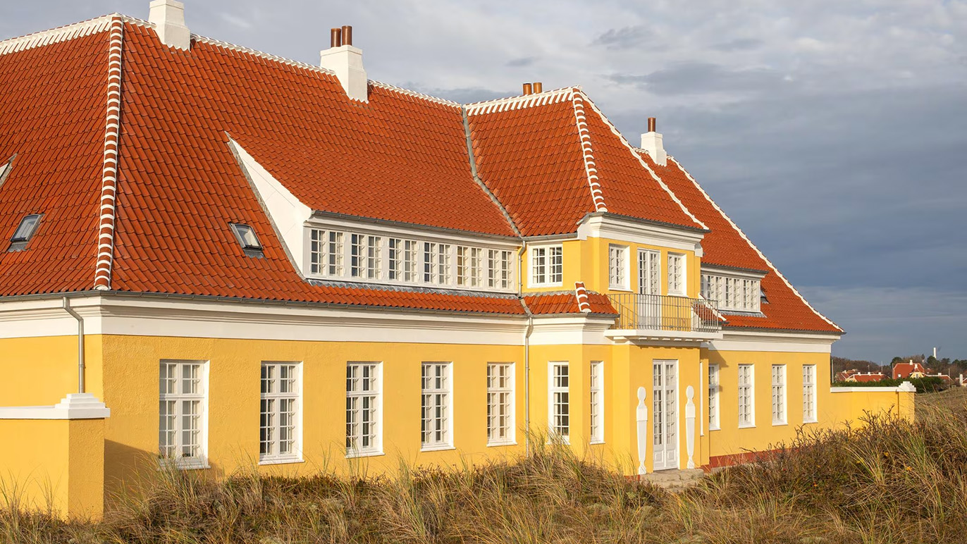 Et stort gult hus med rødt tag og mange vinduer, omgivet af græs og sand.