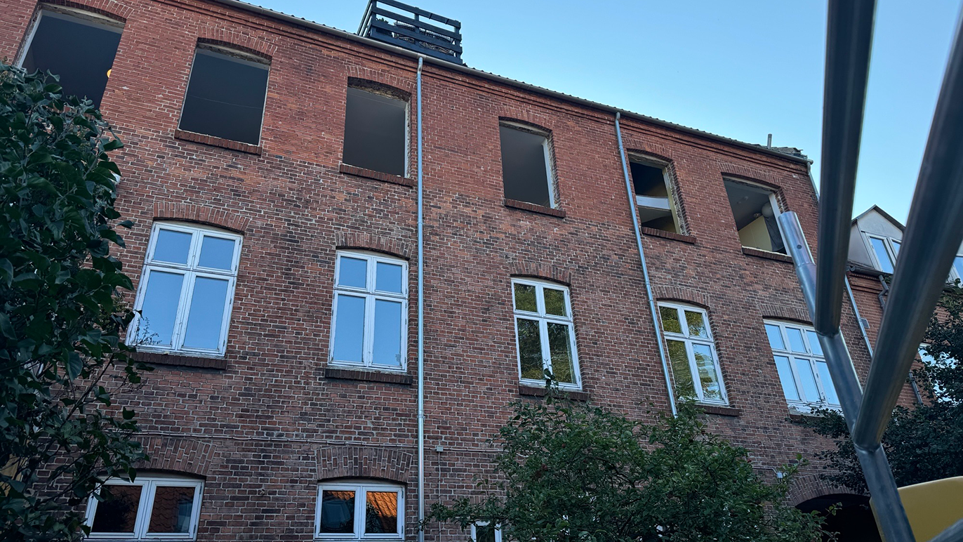 En murstensbygning med flere vinduer og en klar blå himmel i baggrunden. Træer ses i forgrunden.