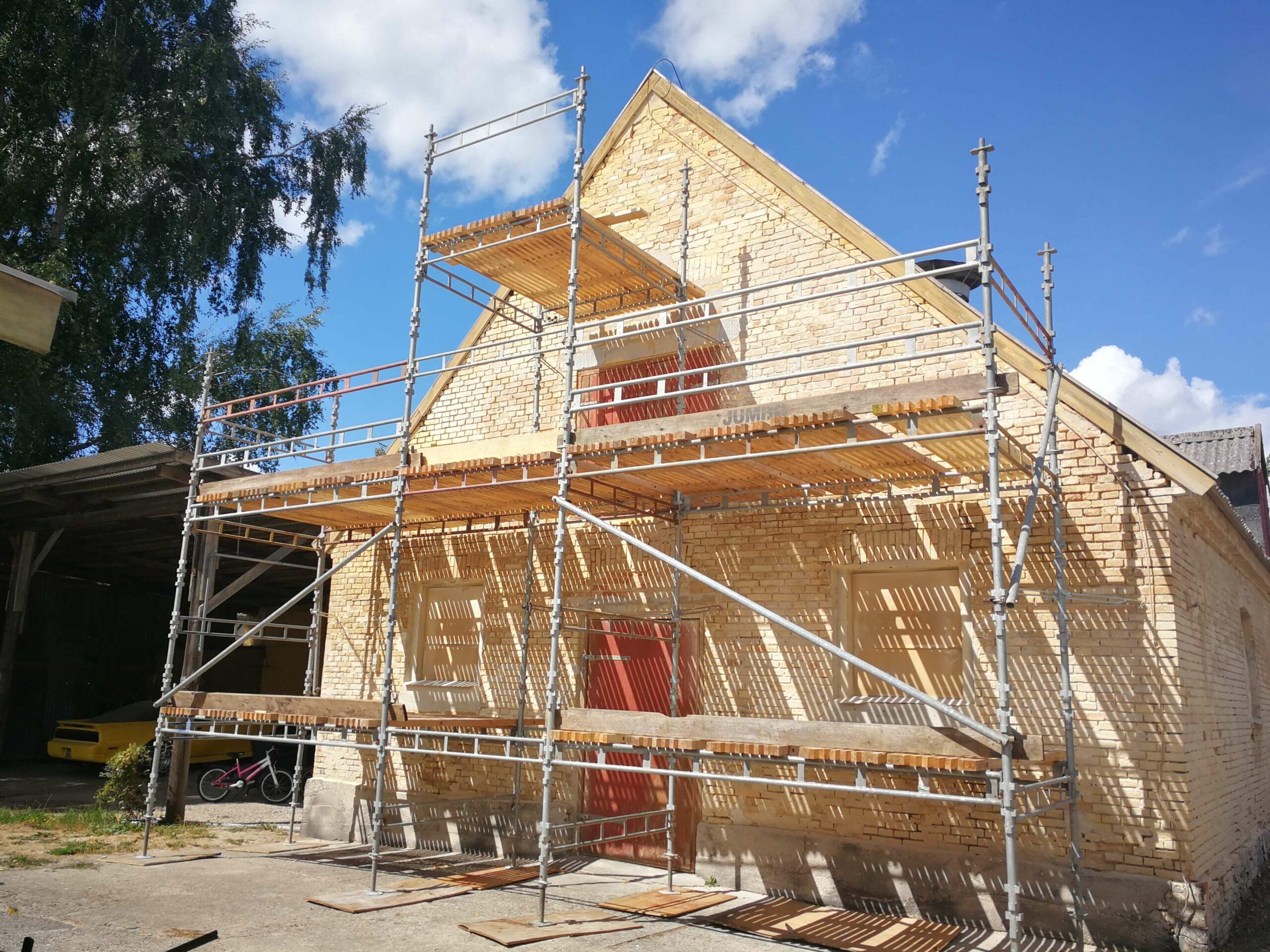 Et hus under konstruktion med stillads omkring, beliggende i en gård med blå himmel og skyer.