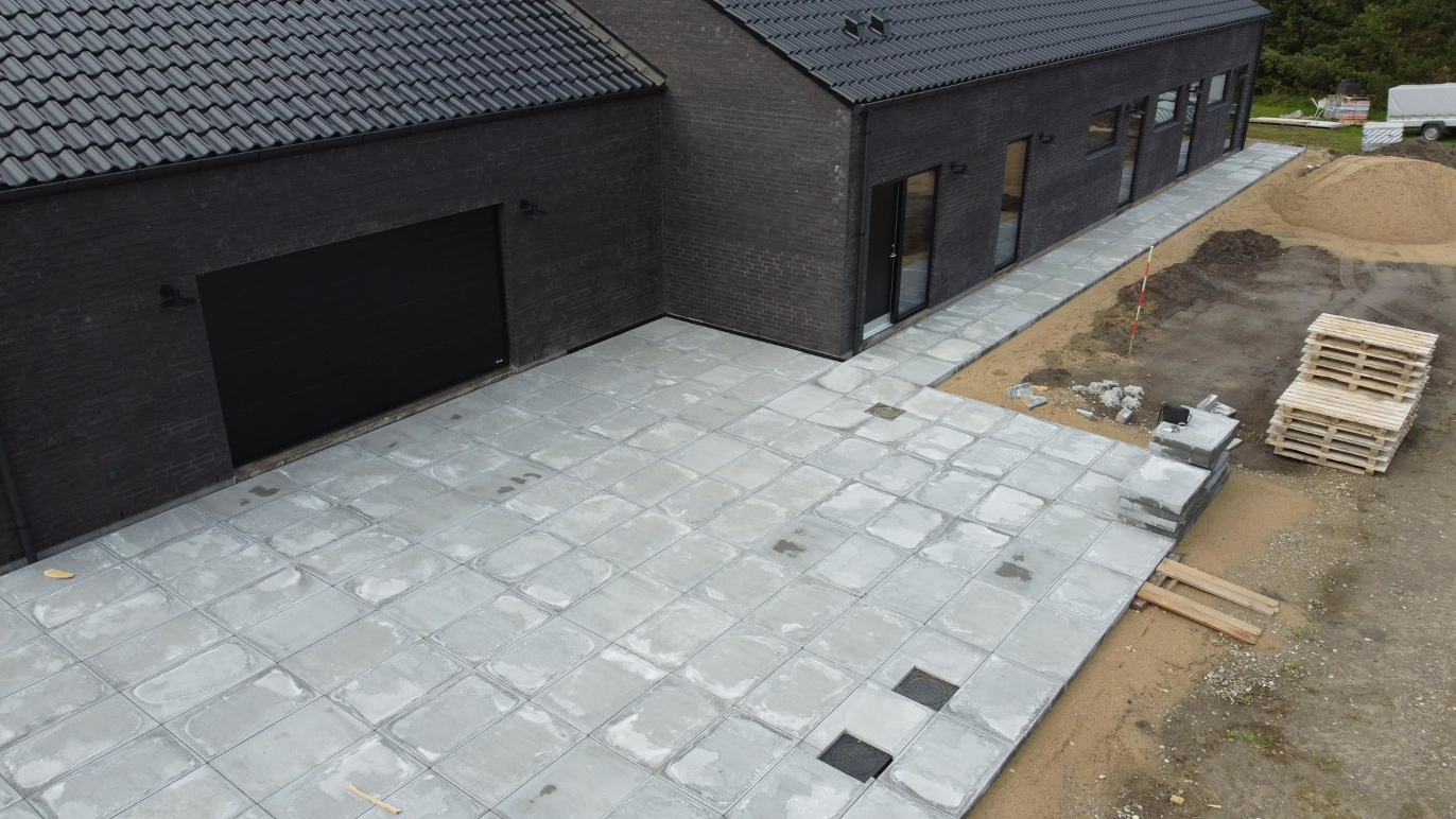 Nybelagt flisebelægning ved en moderne sort bygning med garage og byggeplads i baggrunden.