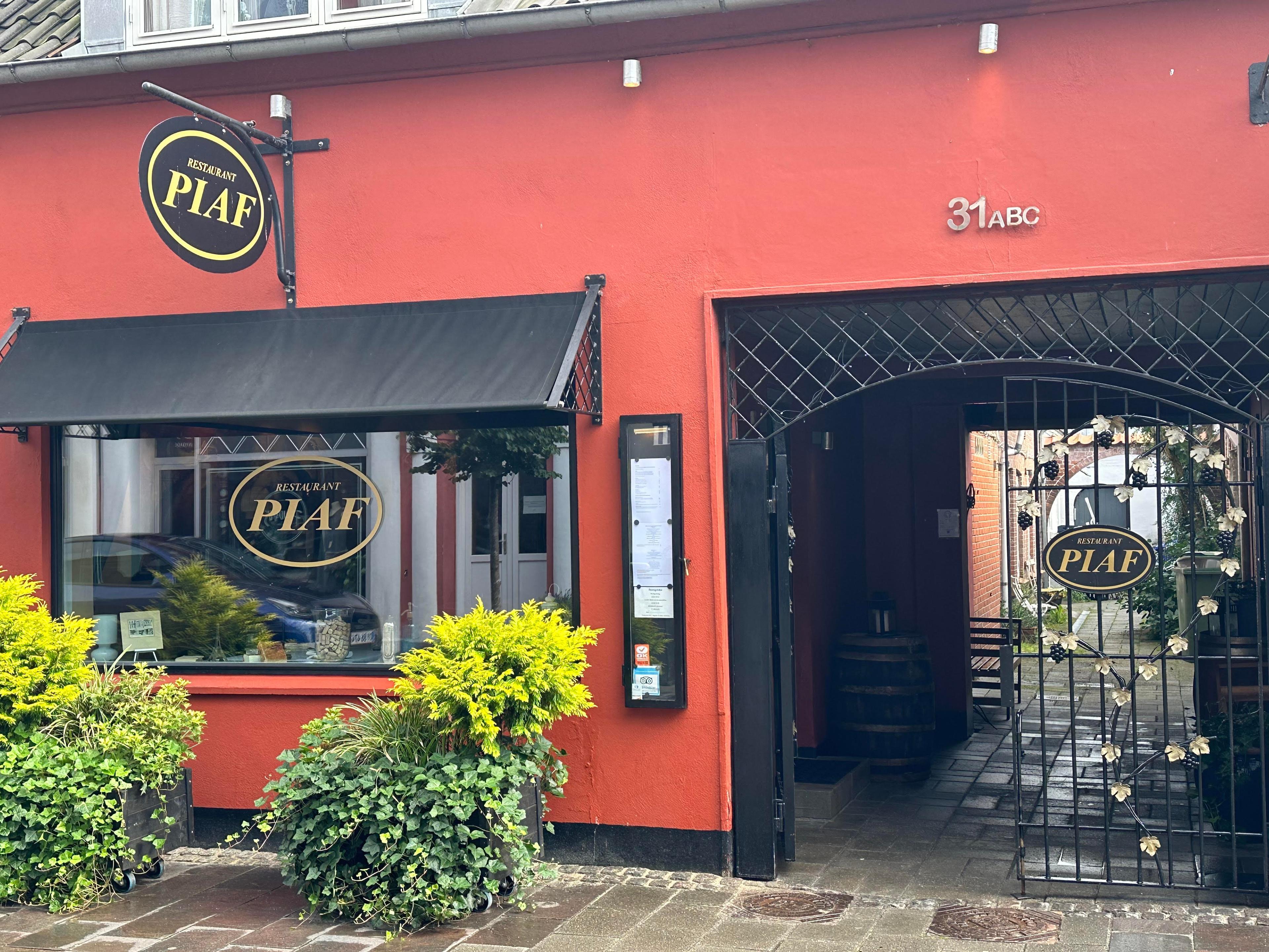 Restaurant Piaf med rød facade, vinduer med sorte markiser og en indgang med gitterport.