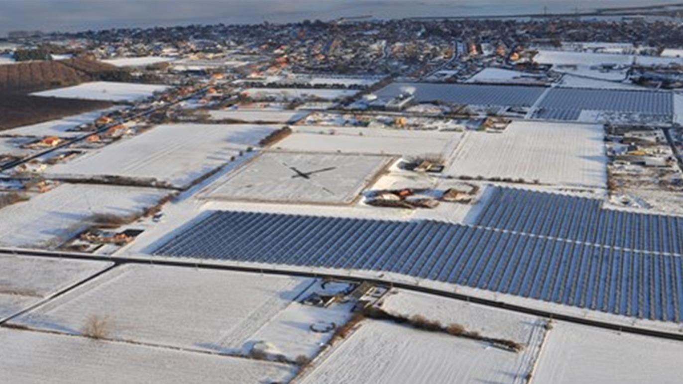 Luftfoto af et snedækket landskab med solcelleanlæg og bygninger i baggrunden.