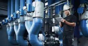 En tekniker i hjelm står ved store blå rør og kontrollerer data på en tablet i en industriel installation.