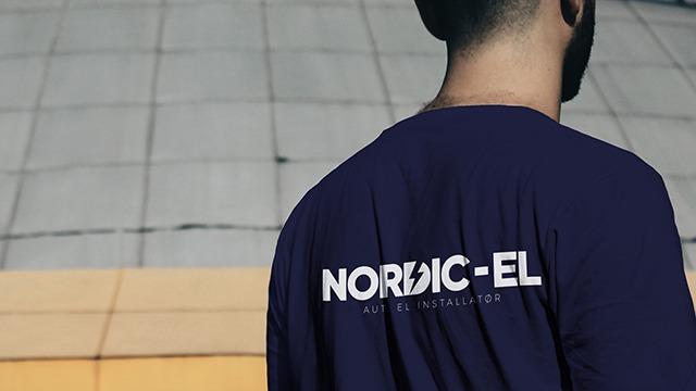 Mand iført en mørkeblå t-shirt med teksten "NORDIC-EL" på ryggen, stående foran en bygning.