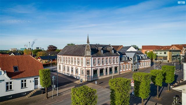 En smuk bygning med spidst tag og grønne træer langs vejen i en by med blå himmel og sollys.