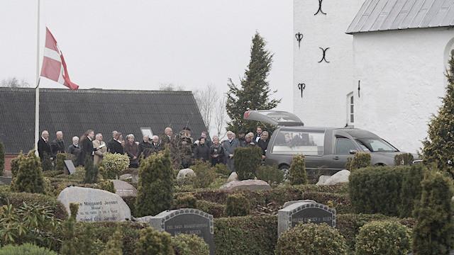 Begravelse ved en kirke med sørgende mennesker og en bil i en gravplads omgivet af gravsten.