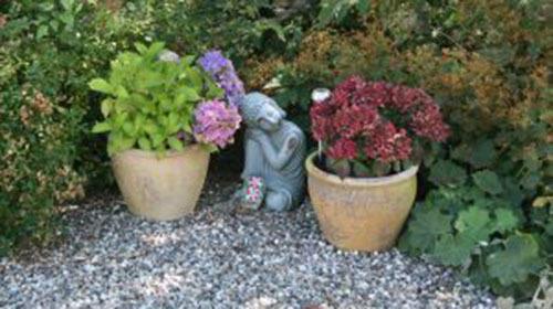 To potter med blomster og en statue af en dreng, der sidder blandt grønne planter i en have.