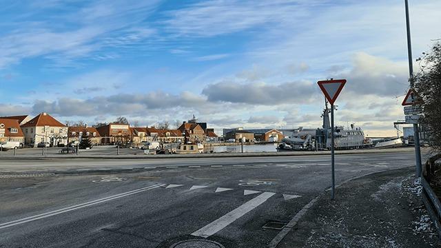 En havn med både, bygninger og en blå himmel med skyer i baggrunden. Vejen fører til vandet.