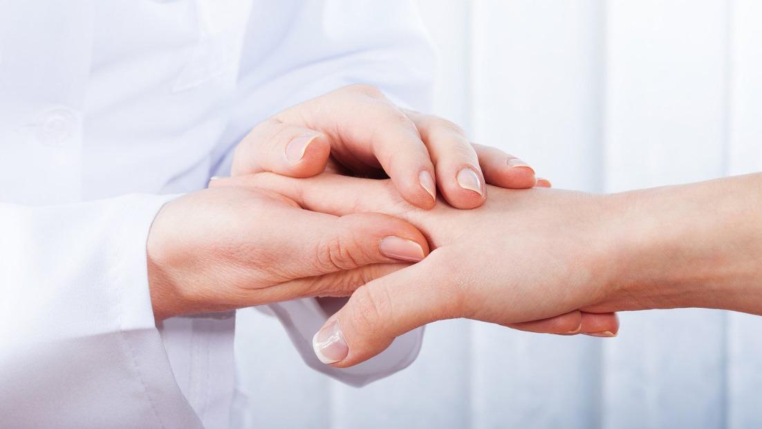 Hænder fra en læge, der undersøger en patients hånd i et klinisk miljø.