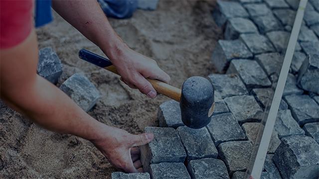 Hånd, der holder en hammer, mens der lægges brosten på en sandbund.