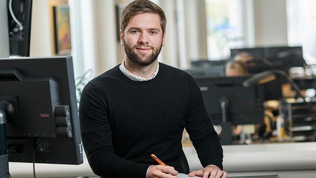 En mand i sort trøje sidder ved et skrivebord med en computer og skriver noter.