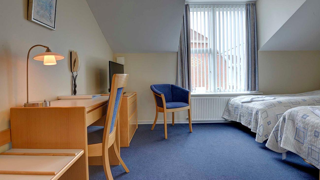 Lyst hotelværelse med to senge, skrivebord, stol og vindue med gardiner.