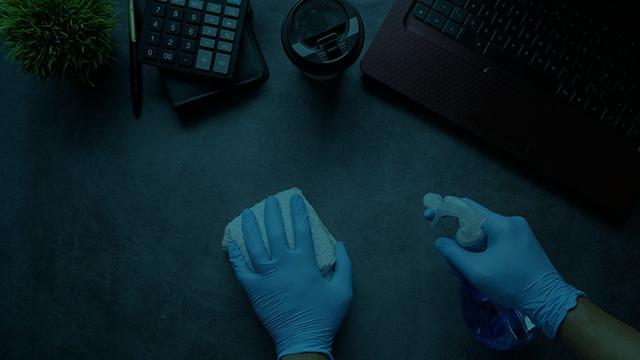 Hånd med gummihandsker, der rengør en overflade med sprayflaske og klud ved en computer og en kop kaffe.