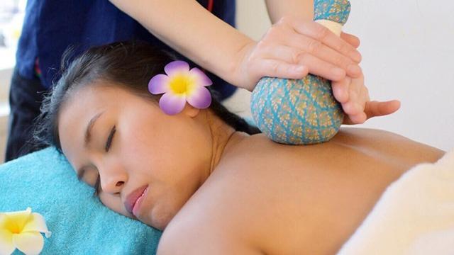 En kvinde får en afslappende massage med en urtepose, mens hun ligger på en blå håndklæde med blomster.