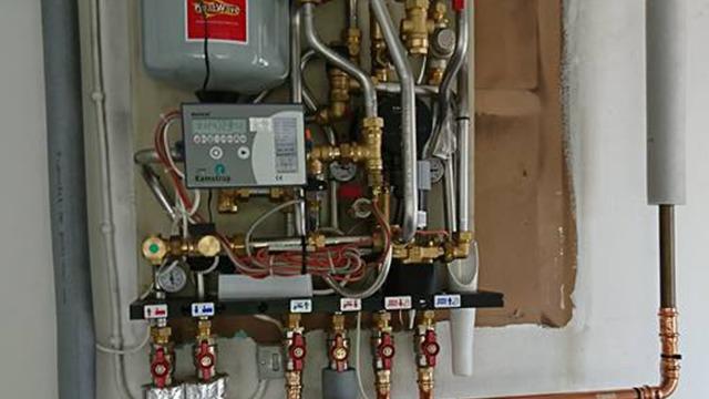 Et komplekst varmesystem med rør, ventiler og en varmepumpe monteret på en væg.