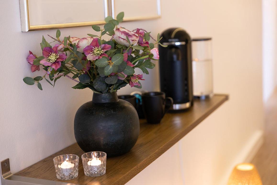 En hylde med en vase med blomster, en kaffemaskine, kopper og stearinlys i et moderne rum.