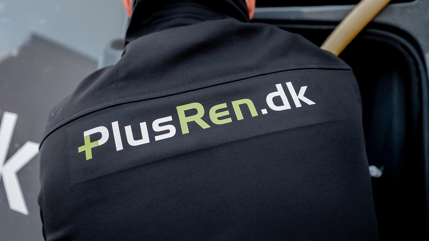 En person iført en sort jakke med logoet PlusRen.dk, der arbejder på en bil.