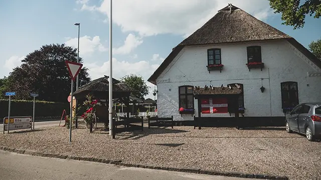 Et hvidt hus med stråtag og blomsterkasser, beliggende ved en vej med trafikskilt og parkering.