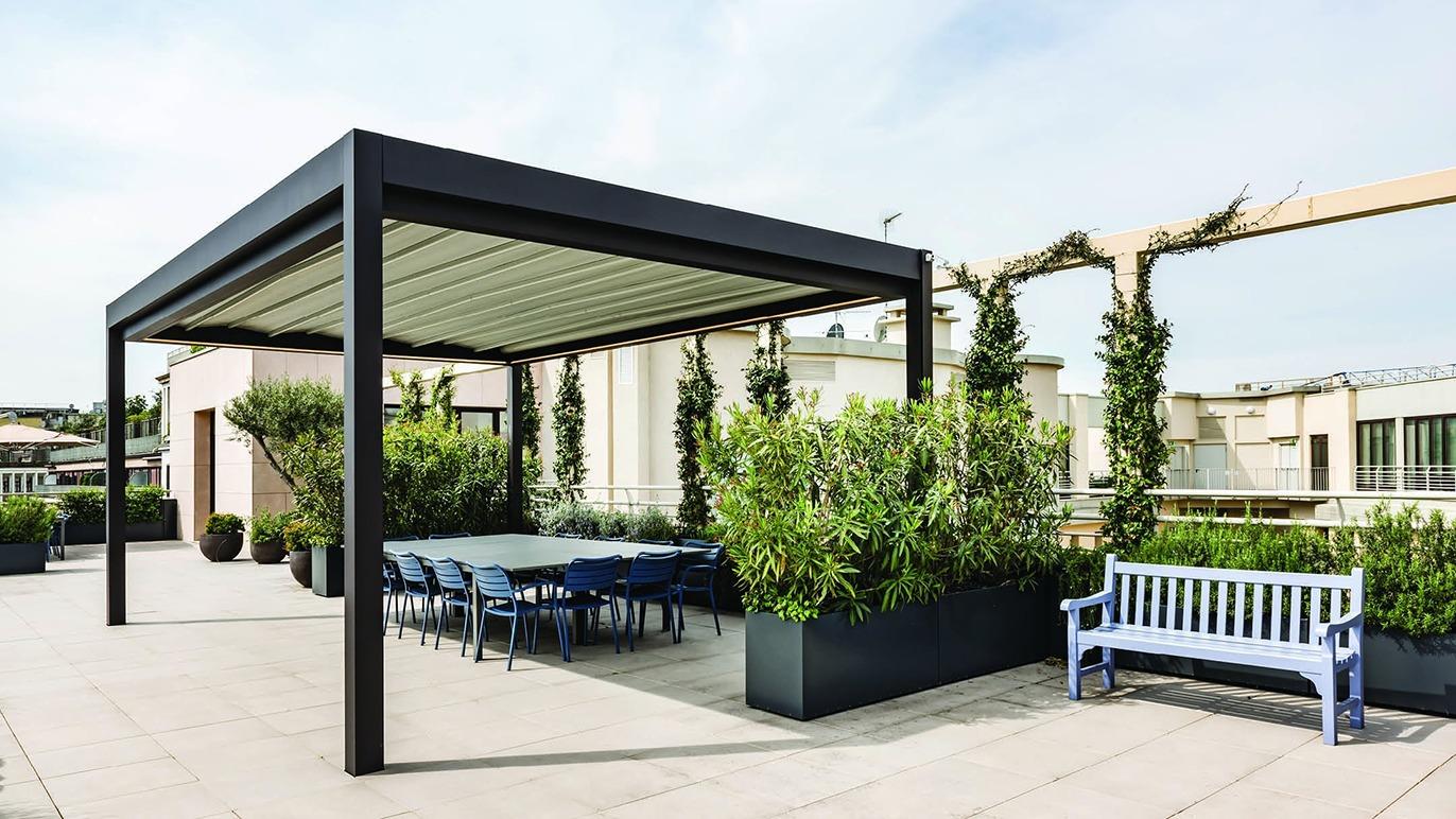 Moderne tagterrasse med pergola, grønne planter og et stort spisebord under en klar himmel.