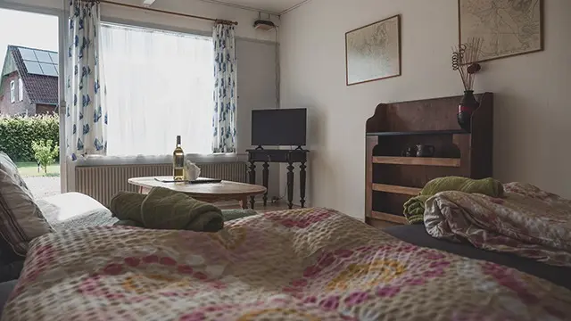 Et hyggeligt værelse med en seng, et bord med vinflaske, en tv og vindue med udsigt til haven.