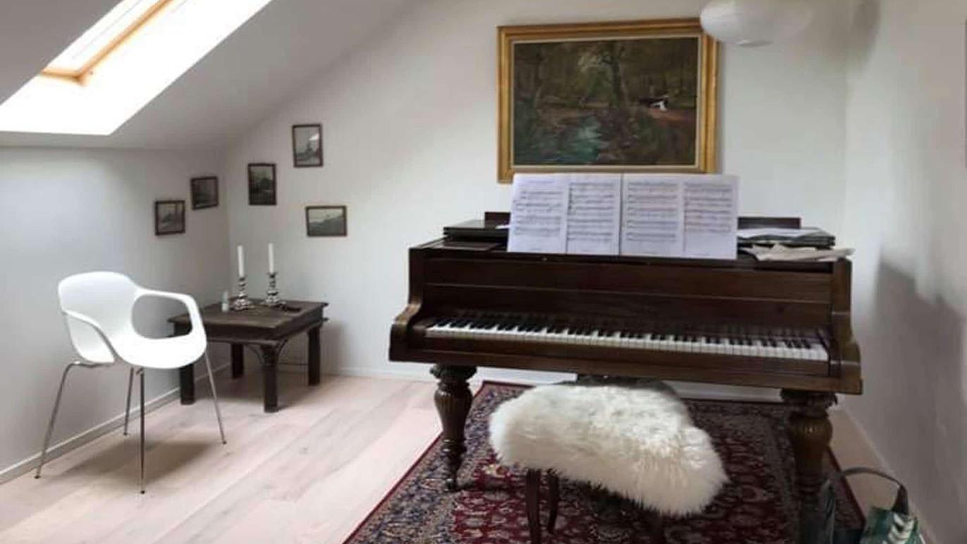 Et klassisk klaver med noder står i et lyst rum med billeder på væggene og en hvid stol ved siden af.