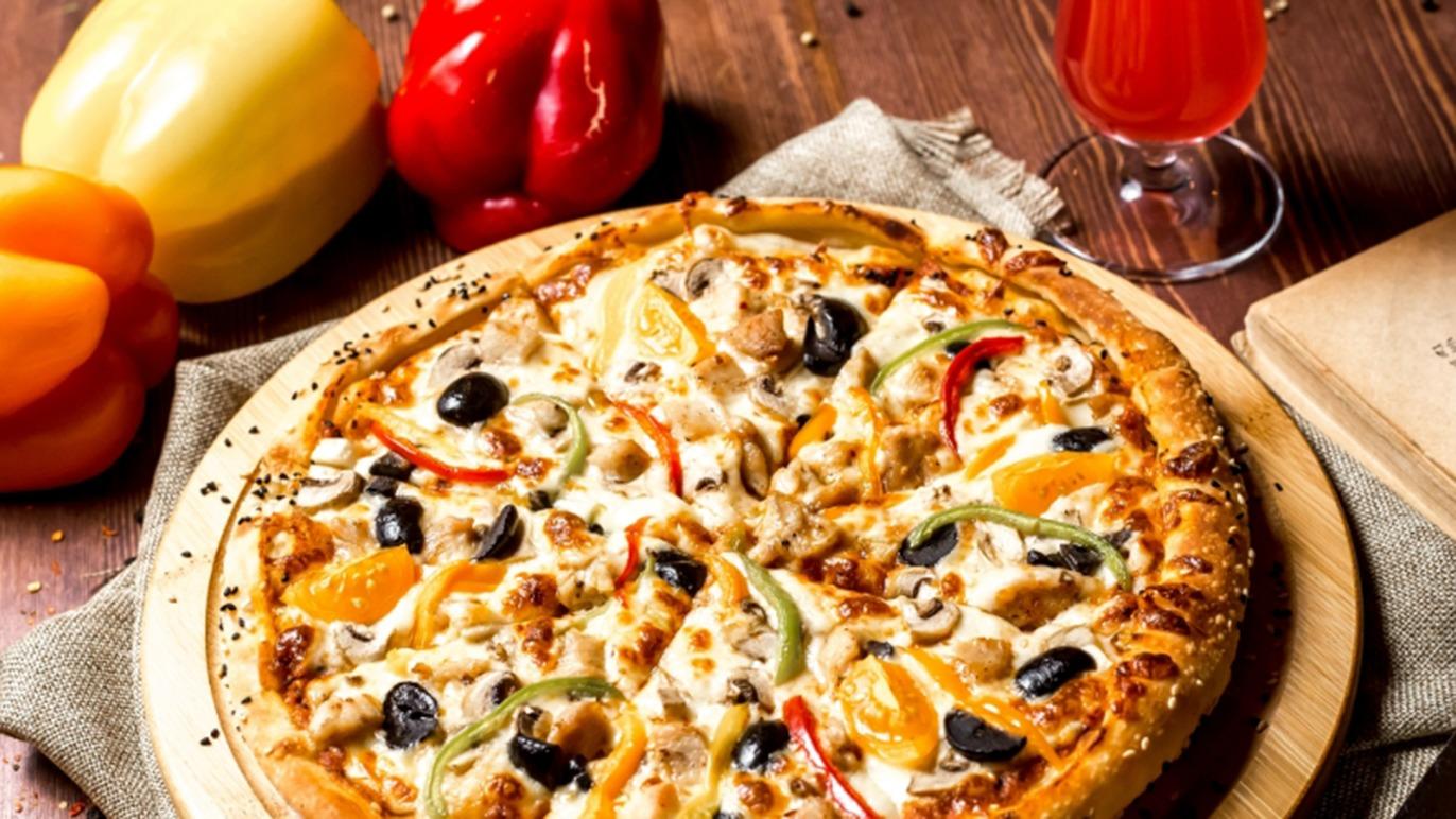 En lækker pizza med skinke, ost, grøntsager og oliven, serveret med en drink og farverige peberfrugter i baggrunden.