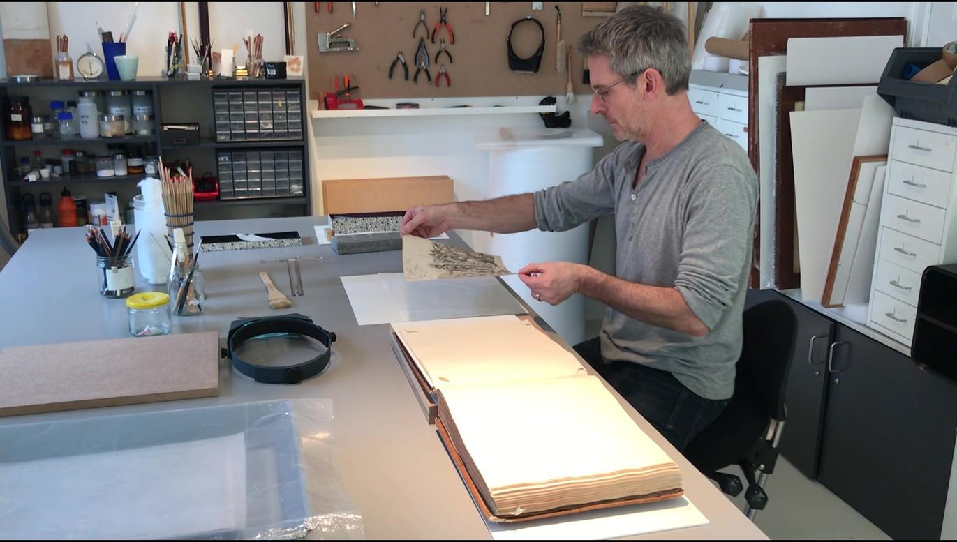En mand sidder ved et bord i et værksted og arbejder med papir og værktøj til kunstneriske projekter.