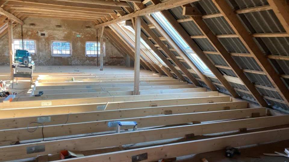 Loftrum med synlige træbjælker og gulvplanker, der venter på installation af isolering og gulvbelægning.