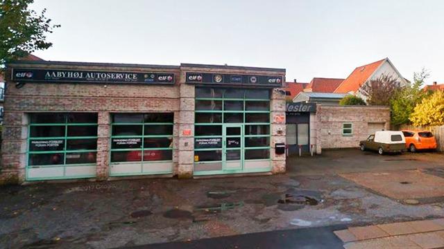 Autoværksted med store vinduer og flere biler parkeret foran bygningen på en solrig dag.