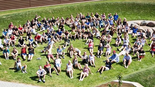 En stor gruppe mennesker sidder på græsset i solen, iført sommertøj og smilende mod kameraet.