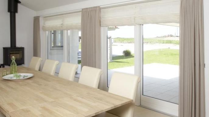 Moderne spisestue med et stort træbord, beige stole og udsigt til haven gennem glasdøre.