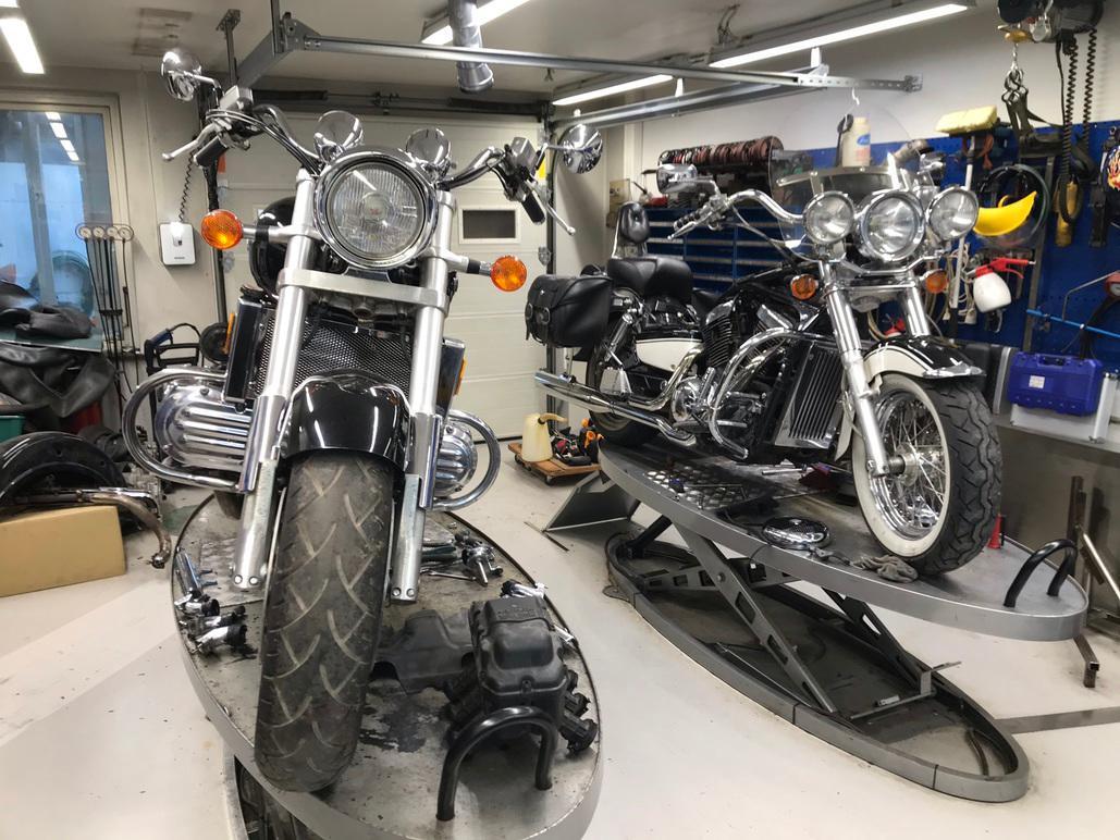 To motorcykler i et værksted, den ene med kromdetaljer og den anden med sort læder, står på en løft.
