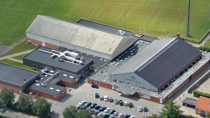 Luftfoto af en stor sportsfacilitet med flere bygninger og parkerede biler på en græsplæne.