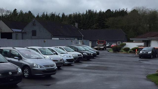 En parkeringsplads med flere grå og sølvfarvede biler, omgivet af bygninger og træer i baggrunden.