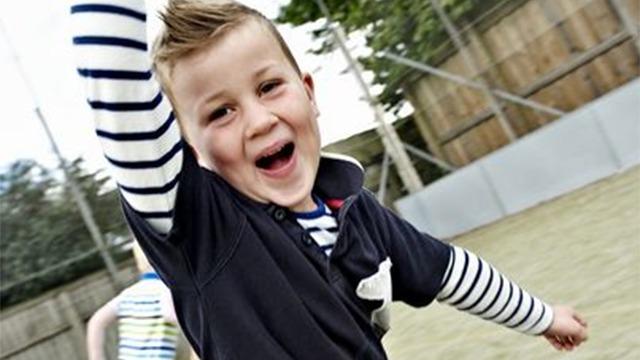 Dreng med kort hår og stribet trøje, der smiler og vinker på en legeplads.