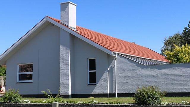Et moderne hus med rød tag og hvid mur, beliggende i et grønt område under en klar blå himmel.
