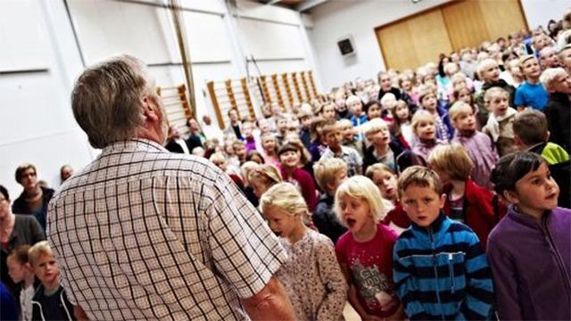 En stor gruppe børn synger sammen, mens en voksen står foran dem i en hal.