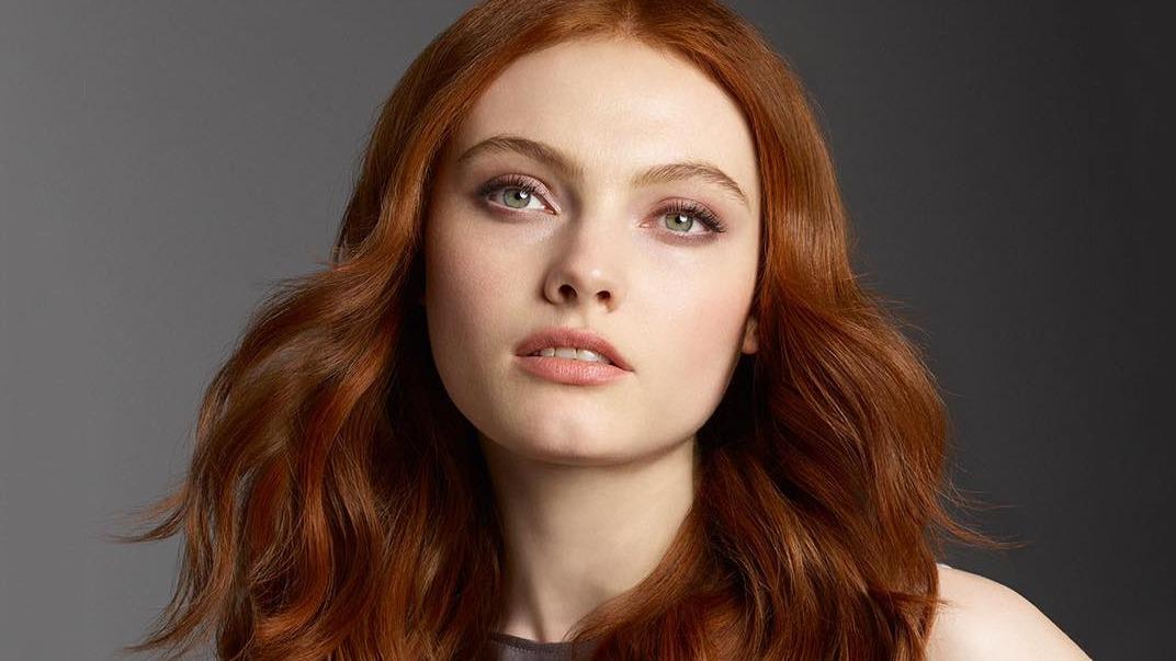 Portræt af en ung kvinde med langt, bølget rødt hår og naturlig makeup, der ser direkte ind i kameraet.