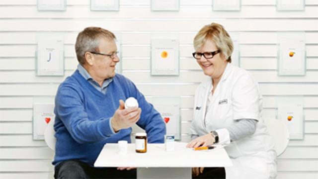 En ældre mand og en kvinde i hvid kittel sidder ved et bord og diskuterer medicin i et lyst rum.