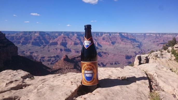 En flaske øl står på en klippe med Grand Canyon i baggrunden under en klar blå himmel.