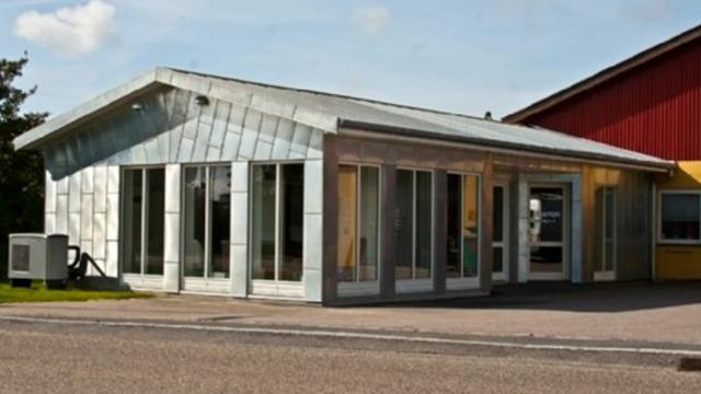 Moderne bygning med store vinduer og metaltag, beliggende ved vejen. Rød bygning i baggrunden.