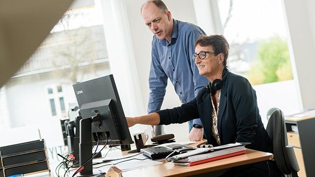 To personer arbejder sammen ved et skrivebord i et moderne kontormiljø, med en computer og kontorudstyr.