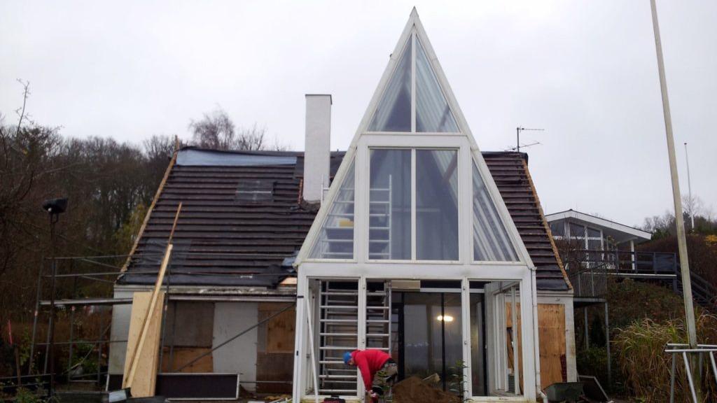 Moderne hus med spids tag og store vinduer, under konstruktion med stillads og håndværker i rødt tøj.