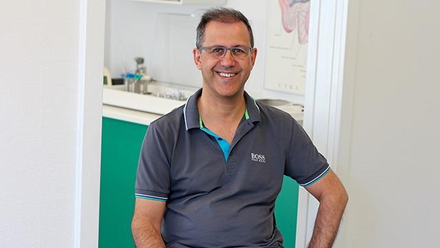En smilende mand i briller sidder i en klinik med grøn væg og medicinsk udstyr i baggrunden.